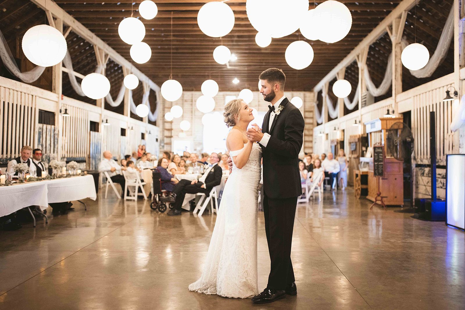Bride and groom share a first dance under white paper lanterns in the barn