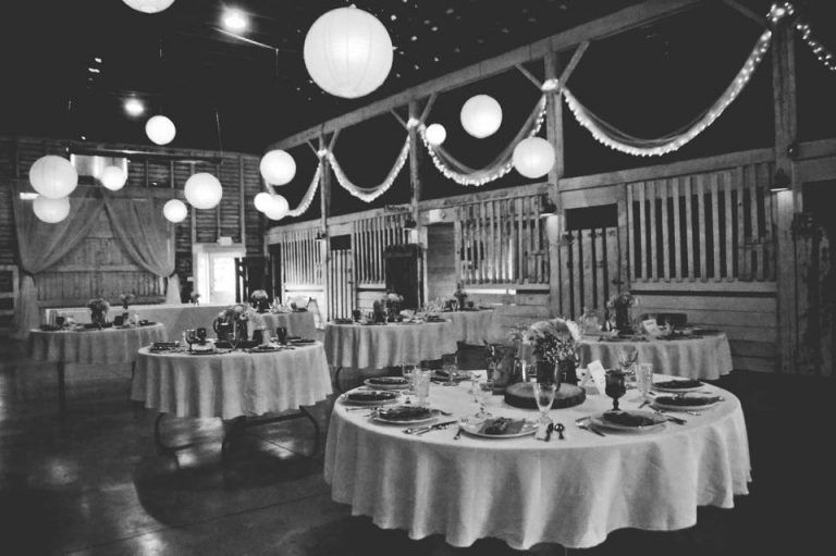 Black and white view of reception tables set beneath lanterns in the barn