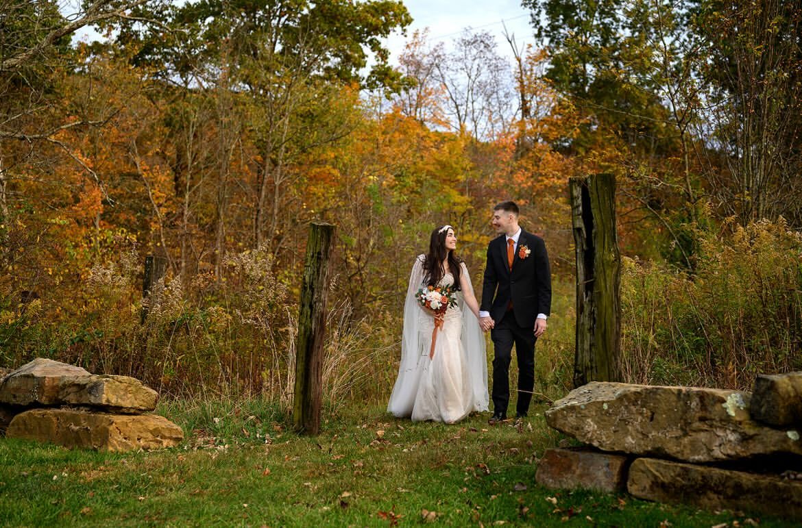 Bride and groom walk hand in hand through an old fence line among autumn trees