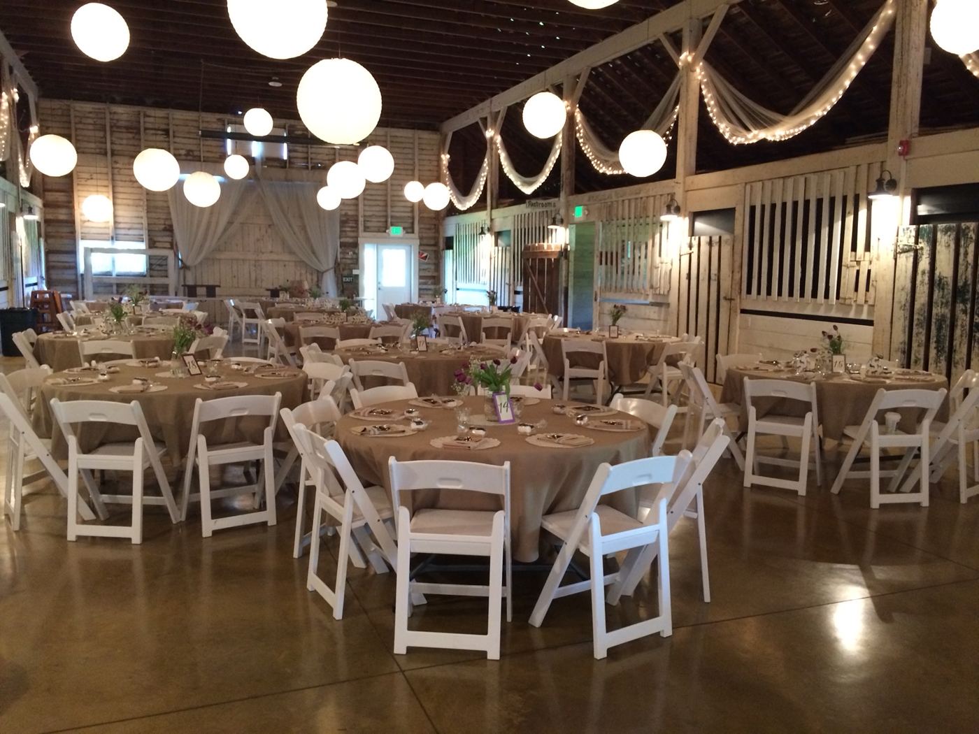 Barn set for a reception with round tables, white chairs and paper lanterns